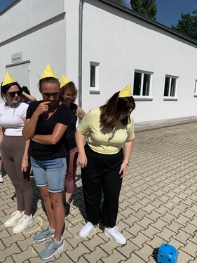 Vier Personen mit gelben Hütchen stehen auf vier gelben Kreisen und würfeln mit einem blauen Würfel.